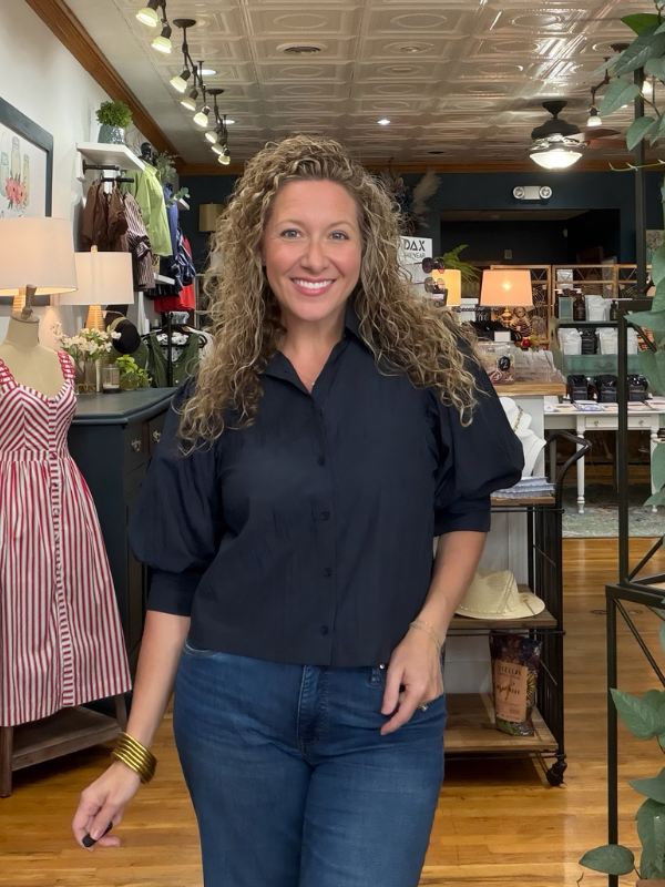 A smiling woman with curly blonde hair stands in a boutique shop wearing a dark navy blue button-down shirt with puff sleeves and blue jeans.