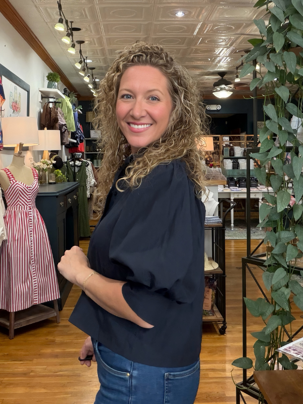 A smiling woman with curly blonde hair wearing a dark navy blue blouse and blue jeans, standing inside a boutique store with wood floors, clothing racks, and decorative plants.