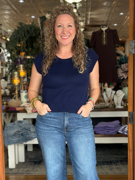 Woman wearing a navy blue top and blue jeans standing in a store with various items in the background.