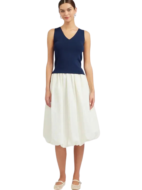 A woman modeling a sleeveless midi dress with a navy blue V-neck top and a flowing off-white skirt, paired with beige flat shoes, against a plain white background.
