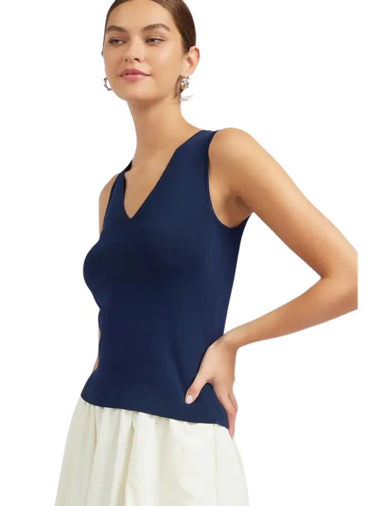 A woman wearing a sleeveless navy blue V-neck top and a white skirt, photographed against a plain white background.