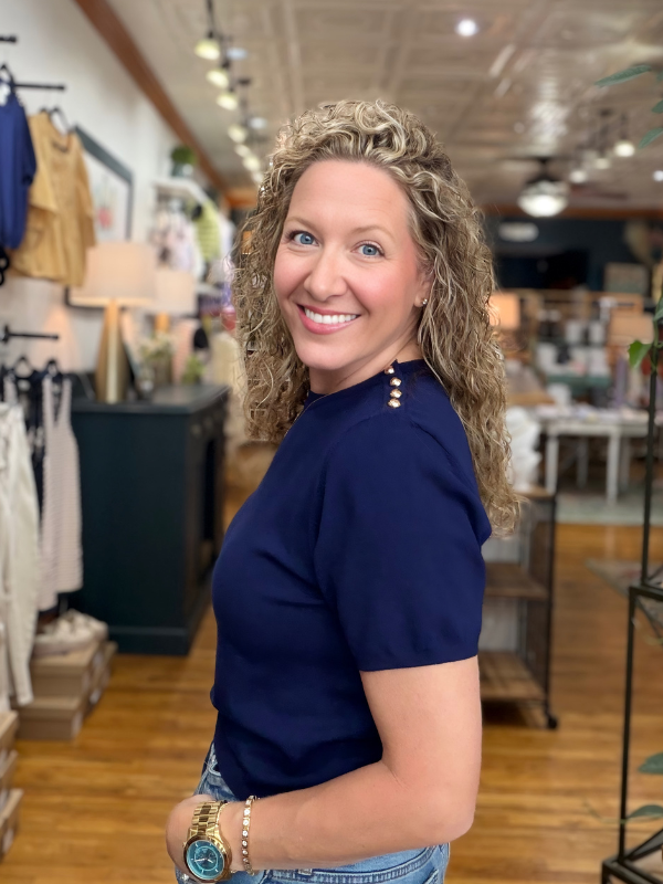 Woman in a navy blue shirt standing in a store with clothing racks and displays in the background.