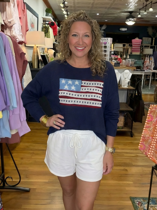 Woman wearing a navy sweater with an American flag design and white shorts in a store setting.