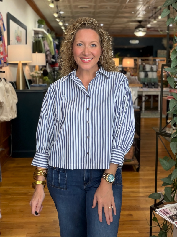 A woman with curly blonde hair stands in a retail store wearing a blue and white vertically striped button-down shirt and dark blue jeans.