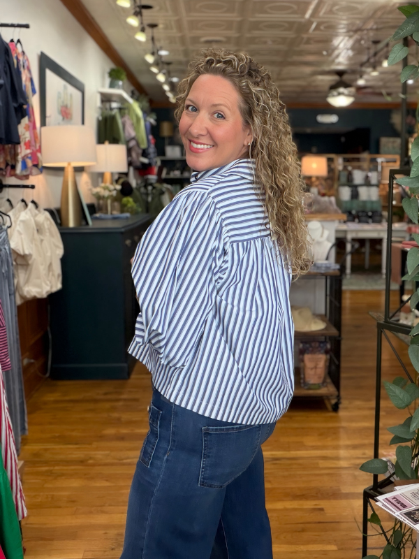 A woman with curly blonde hair wearing a blue and white striped button-up shirt and dark blue jeans, standing in a retail store and looking back over her shoulder.
