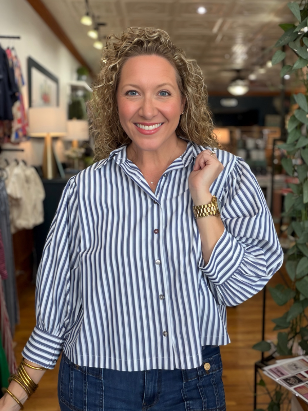 A smiling woman with curly blonde hair wearing a blue and white vertically striped button-down shirt and dark blue jeans, standing inside a boutique store.