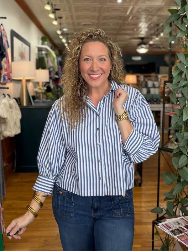 A smiling woman with curly blonde hair stands in a retail store wearing a blue and white vertically striped button-down shirt and dark blue jeans.