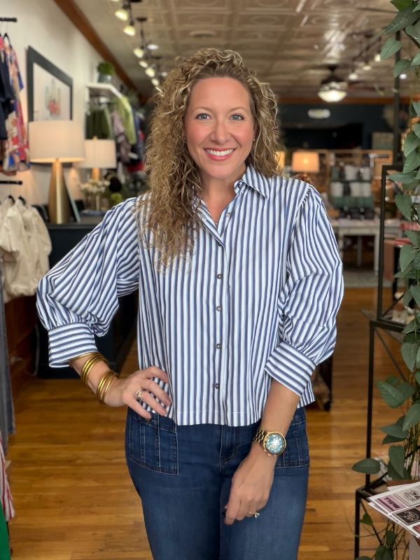 A smiling woman with curly blonde hair stands in a boutique wearing a blue and white vertically striped button-down shirt with puff sleeves, paired with dark blue jeans.