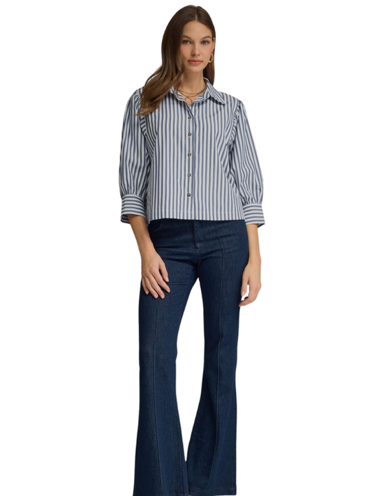 A woman modeling a blue and white vertically striped button-down shirt with three-quarter sleeves, paired with dark blue flare jeans, against a plain white background.