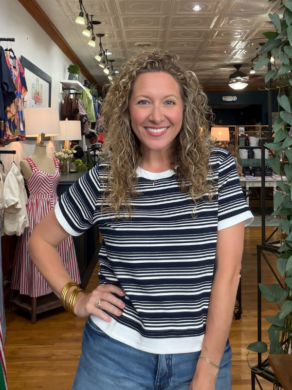 A smiling woman with curly blonde hair stands inside a clothing store wearing a navy and white horizontally striped short-sleeve t-shirt with white ruffle trim, paired with blue jeans.