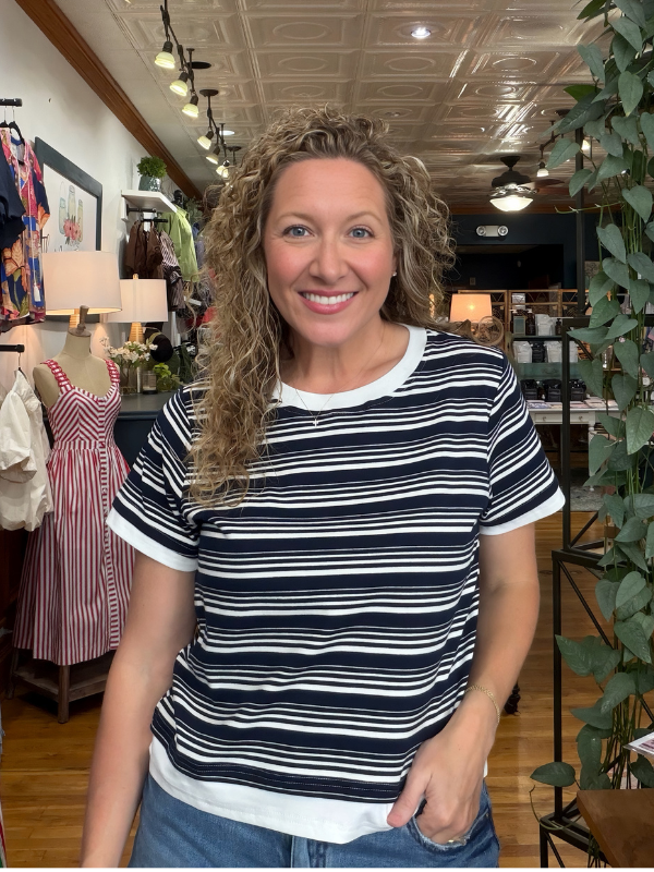 A smiling woman with curly blonde hair wearing a navy and white horizontally striped short-sleeve t-shirt with white ribbed collar, cuffs, and hem, standing in a boutique.