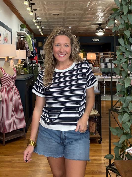 A woman with curly blonde hair stands in a retail store wearing a navy and white horizontally striped short-sleeve t-shirt with white ruffle trim, paired with denim shorts.