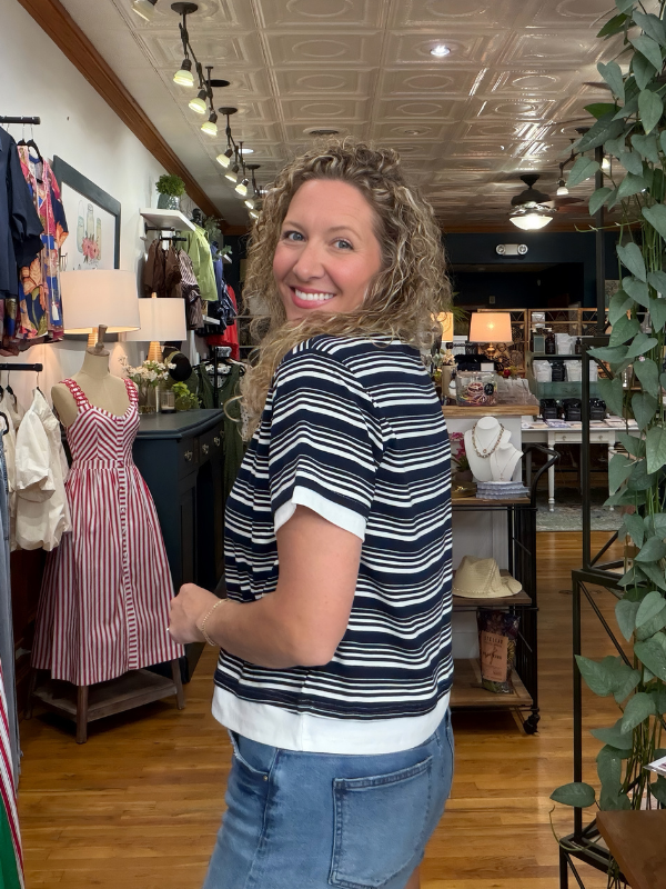 A woman with curly blonde hair stands in a boutique shop, looking back over her shoulder while wearing a navy and white horizontally striped short-sleeve top with a white hem, paired with blue jeans.