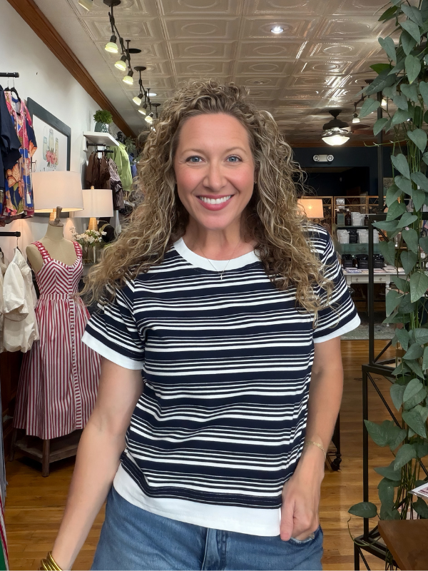 A smiling woman with curly blonde hair wearing a navy and white horizontally striped t-shirt with white ringer trim, standing inside a retail store.
