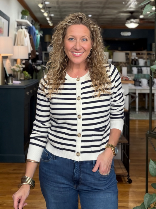 A smiling woman with curly blonde hair stands in a boutique wearing a cream and navy blue horizontally striped button-up cardigan with gold-tone buttons, paired with blue jeans.