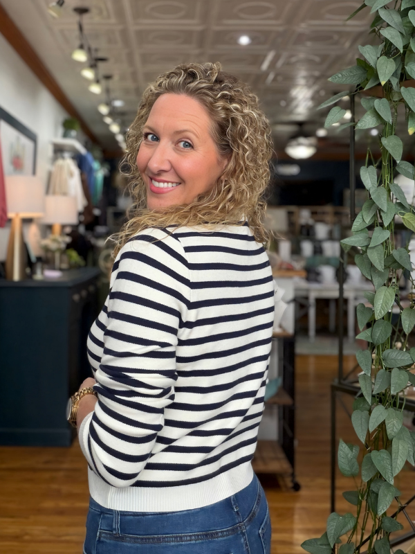 A woman with curly blonde hair is shown from the back, wearing a black and white horizontally striped long-sleeve knit sweater and blue jeans inside a boutique.