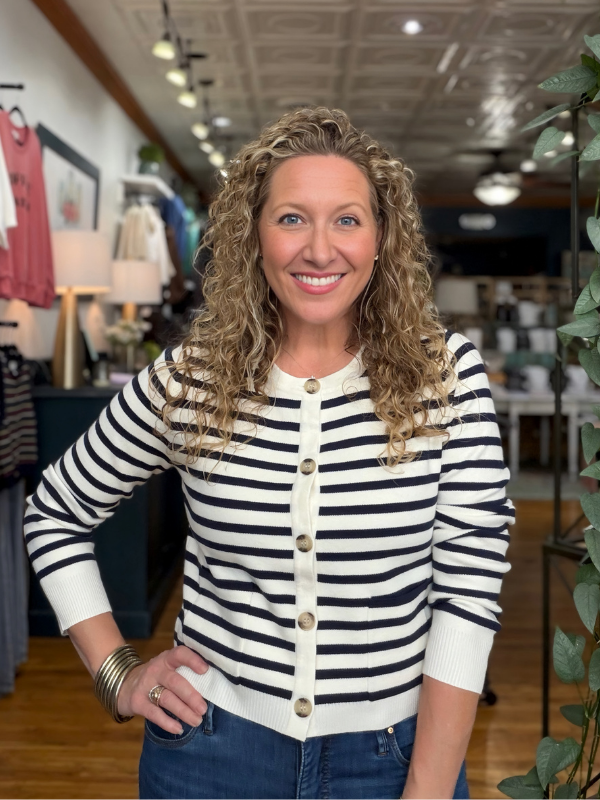 A smiling woman with curly blonde hair stands in a clothing store wearing a navy and white horizontally striped button-front cardigan with three-quarter sleeves and blue jeans.