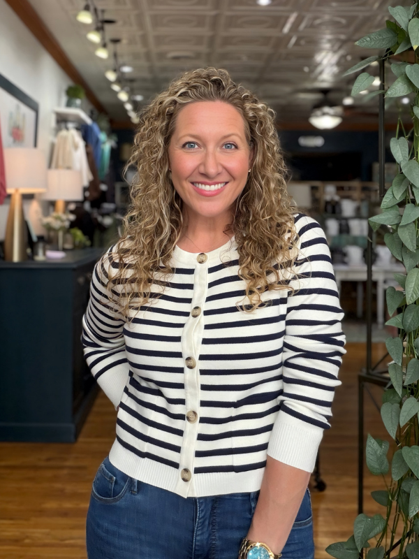 A smiling woman with curly blonde hair wearing a black and white striped button-up cardigan and blue jeans, standing inside a retail store.