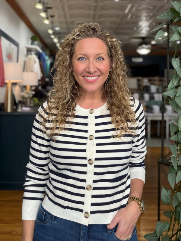 A smiling woman with curly blonde hair wearing a black and white horizontally striped knit cardigan with wooden buttons, paired with blue jeans, standing inside a retail store.