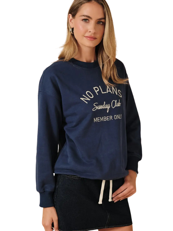 A woman wearing a navy blue crewneck sweatshirt with white text that reads 'NO PLANS Sunday Clubs MEMBER ONLY', paired with a dark denim skirt, against a plain white background.