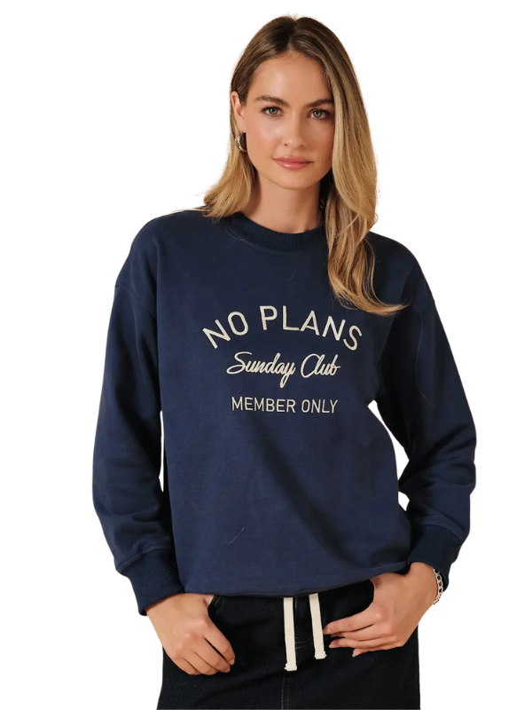 A woman wearing a navy blue crewneck sweatshirt with white text that reads 'NO PLANS Sunday Club MEMBER ONLY', paired with black pants with white drawstrings, against a plain white background.