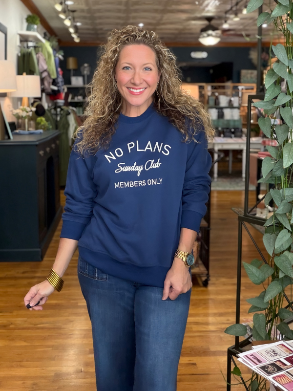 A smiling woman with curly hair stands in a retail store wearing a navy blue crewneck sweatshirt with white text that reads 'NO PLANS Sunday Club MEMBERS ONLY' paired with blue jeans.