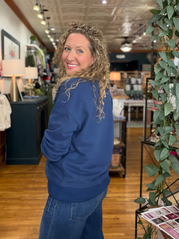 A woman with curly blonde hair smiling and looking back over her shoulder while wearing a royal blue crewneck sweatshirt and blue jeans inside a retail store.