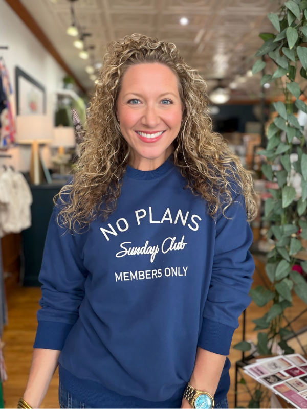 A smiling woman with curly blonde hair wearing a navy blue crewneck sweatshirt with white text that reads 'NO PLANS Sunday Club MEMBERS ONLY', standing in a retail store interior.