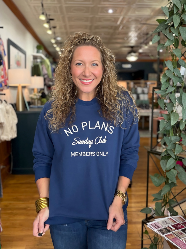 A smiling woman with curly hair stands in a retail store wearing a navy blue crewneck sweatshirt with white text that reads 'NO PLANS Sunday Club MEMBERS ONLY'.