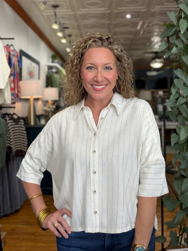 A smiling woman with curly blonde hair wearing a cream-colored, short-sleeve button-up shirt with thin vertical stripes, paired with dark blue jeans, standing inside a boutique.