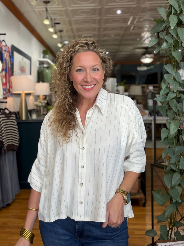 A smiling woman with curly blonde hair stands in a boutique wearing a white button-up shirt with thin vertical pinstripes and dark blue jeans.