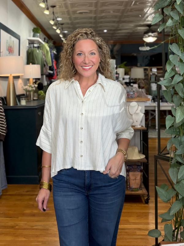 A smiling woman with curly blonde hair stands in a boutique wearing a white button-up shirt with thin vertical pinstripes and dark blue jeans.