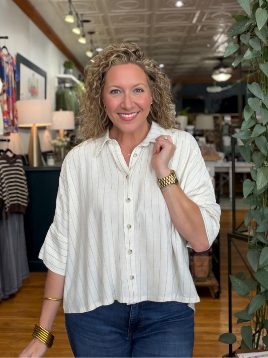 A smiling woman with curly blonde hair stands in a boutique wearing a cream-colored, short-sleeve button-up shirt with thin vertical stripes and dark blue jeans.