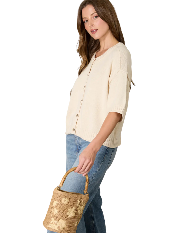 Woman wearing a cream cardigan and blue jeans, holding a woven handbag on a white background