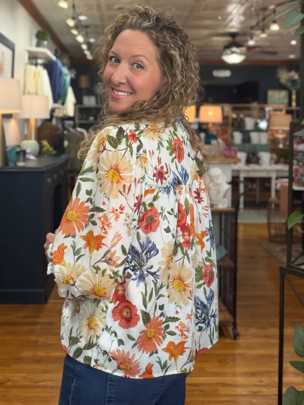 A woman modeling a long-sleeve button-up blouse with a white base and a vibrant multicolor floral print, paired with blue jeans 