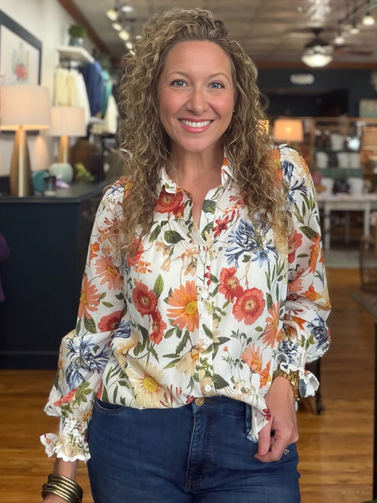 A woman modeling a long-sleeve button-up blouse with a white base and a vibrant multicolor floral print, paired with blue jeans 