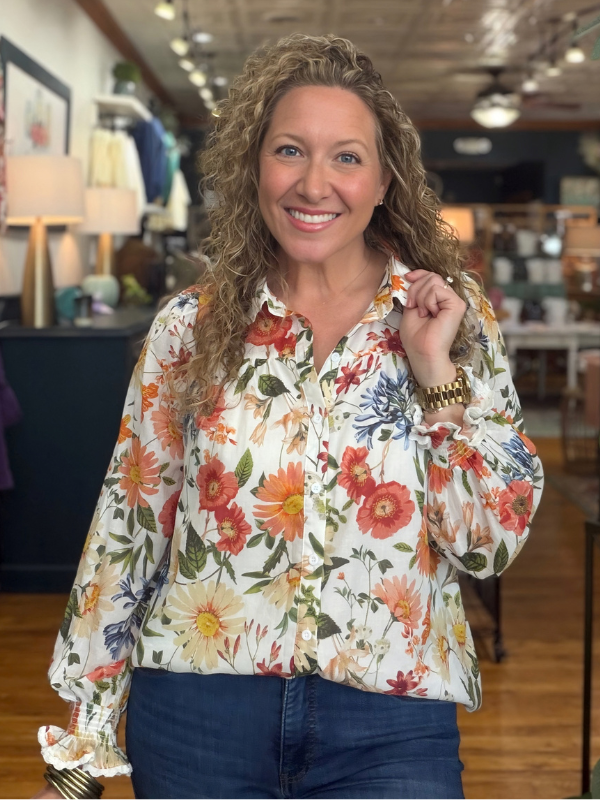 A woman modeling a long-sleeve button-up blouse with a white base and a vibrant multicolor floral print, paired with blue jeans 
