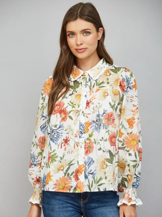 A woman modeling a long-sleeve button-up blouse with a white base and a vibrant multicolor floral print, paired with blue jeans against a solid gray studio background.