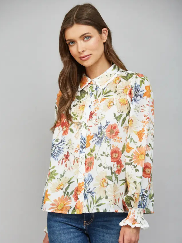 A woman wearing a long-sleeve button-up blouse with a colorful floral print on a cream background, paired with blue jeans, photographed against a solid gray studio background.