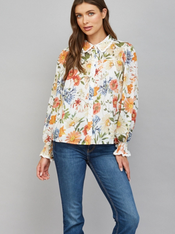 A woman modeling a long-sleeve button-up blouse with a dense multicolor floral print on a cream background, paired with blue jeans, against a solid gray studio background.