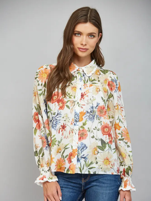 A woman modeling a long-sleeve button-up blouse with a vibrant multicolor floral print on a cream background, paired with blue jeans, against a solid gray studio background.