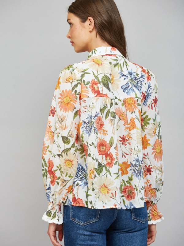 A woman shown from the back wearing a long-sleeve white blouse with a vibrant multicolor floral print and blue jeans, set against a plain gray studio background.