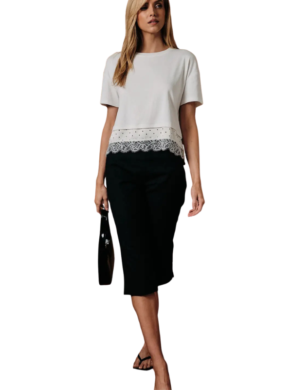 A woman modeling a short-sleeve top with a white upper section and a black lace hem, paired with a black midi skirt and holding a black handbag against a plain white background.