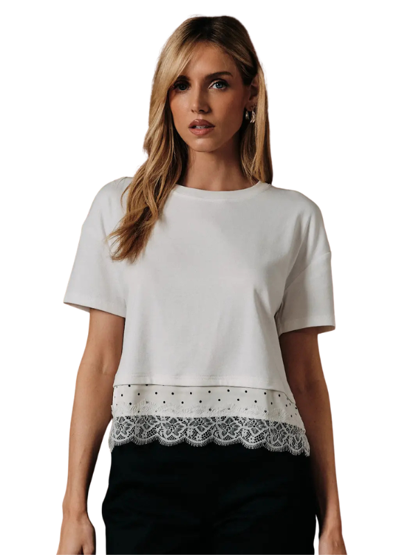 A woman wearing a white short-sleeve crop top with a decorative lace and polka-dot hem, paired with black pants, against a plain white background.