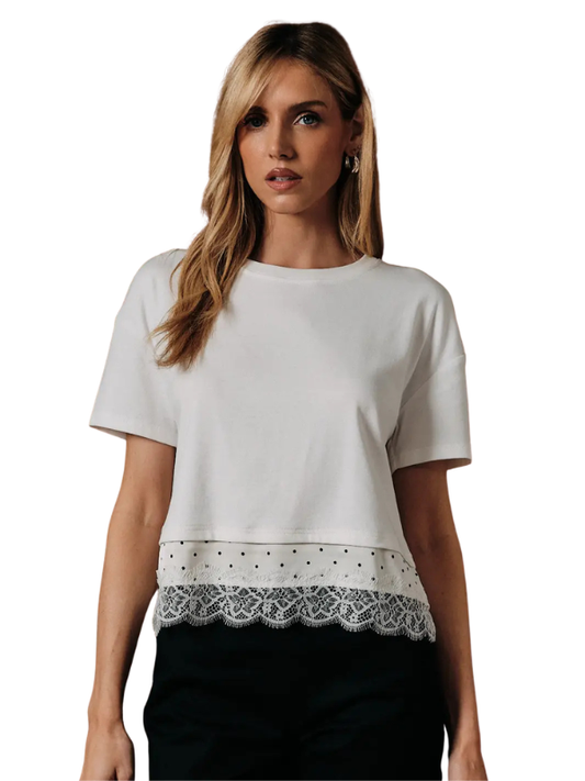 A woman wearing a white short-sleeve crop top with a decorative lace and polka-dot hem, paired with black pants, against a plain white background.