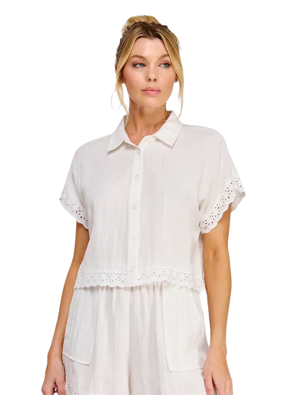 A woman modeling a white short-sleeve button-up shirt with eyelet lace trim on the sleeves and hem, paired with matching white bottoms, against a plain white background.