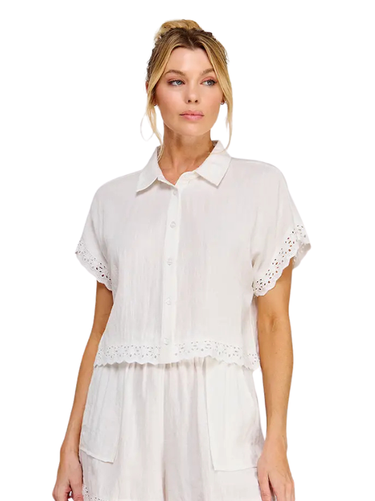 A woman modeling a white short-sleeve button-up shirt with eyelet lace trim on the sleeves and hem, paired with matching white bottoms, against a plain white background.
