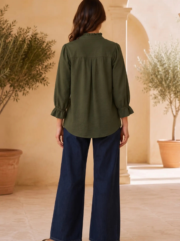 A woman viewed from behind wearing an olive green long-sleeve blouse with ruffled cuffs and dark blue wide-leg pants, standing in a sunlit courtyard with beige walls and potted plants.