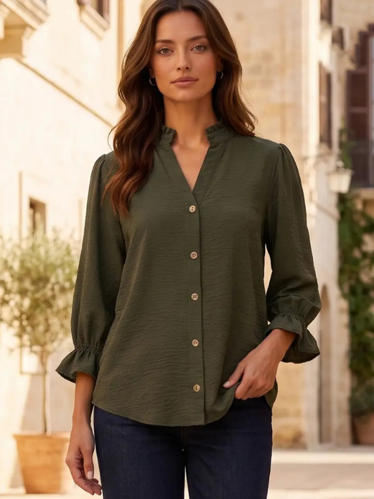 A woman wearing an olive green long-sleeve button-up blouse with a high folded collar and dark blue jeans, standing in an outdoor street setting with warm-toned buildings in the background.