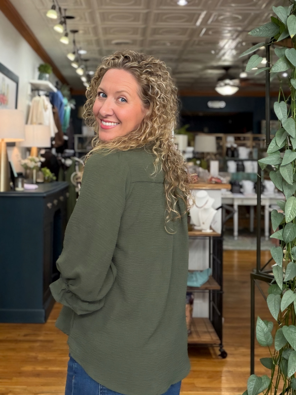 A woman with curly blonde hair wearing an olive green long-sleeve top and blue jeans, smiling and looking back over her shoulder inside a retail store.